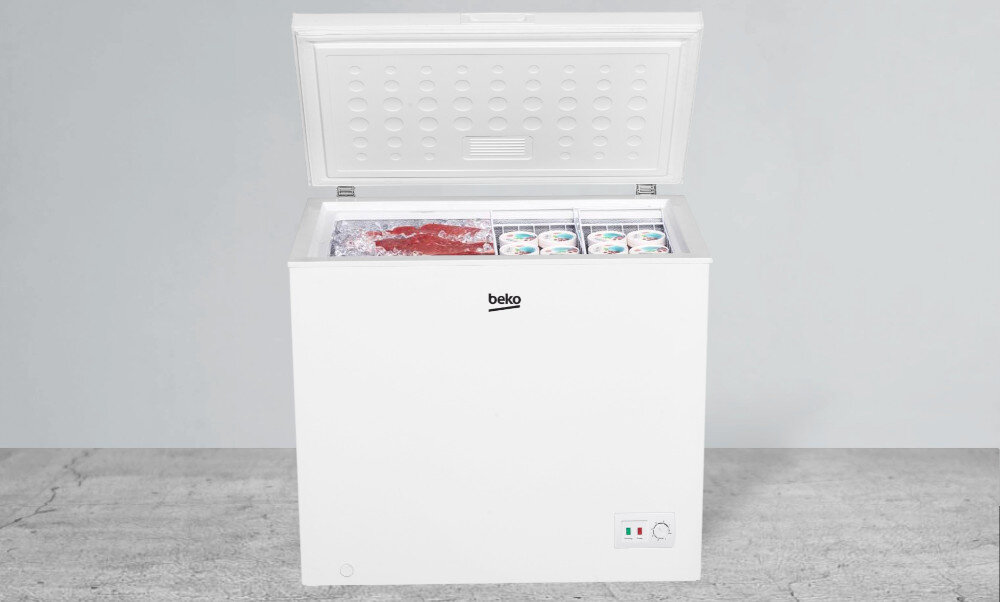 Biała zamrażarka skrzyniowa z otwartą pokrywą, na której widoczne jest logo \'beko\'. Wewnątrz znajdują się różne produkty spożywcze, a na zewnątrz panel z pokrętłem i wskaźnikami.