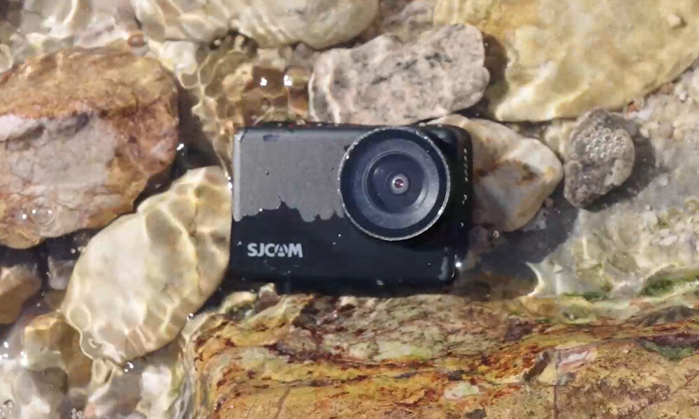 Kamera sportowa SJCAM leży na kamieniach w płytkiej wodzie. Na obudowie widoczne jest logo \'SJCAM\'.