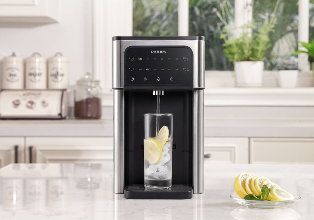 Na blacie kuchennym stoi dyspenser do wody marki PHILIPS, z którego nalewana jest woda do szklanki z lodem i plasterkami cytryny. W tle widoczne są pojemniki na produkty spożywcze oraz rośliny doniczkowe na parapecie.