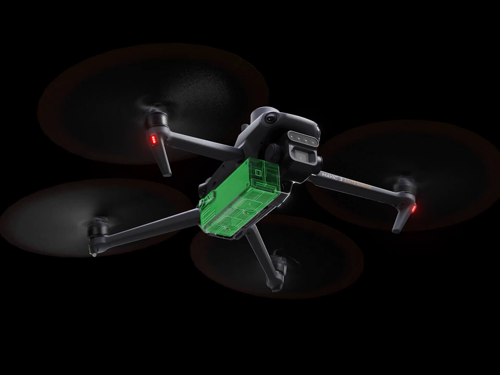 Na obrazie widoczny jest dron z czterema wirnikami, z zielonym elementem na spodzie. Dron unosi się w powietrzu na ciemnym tle.