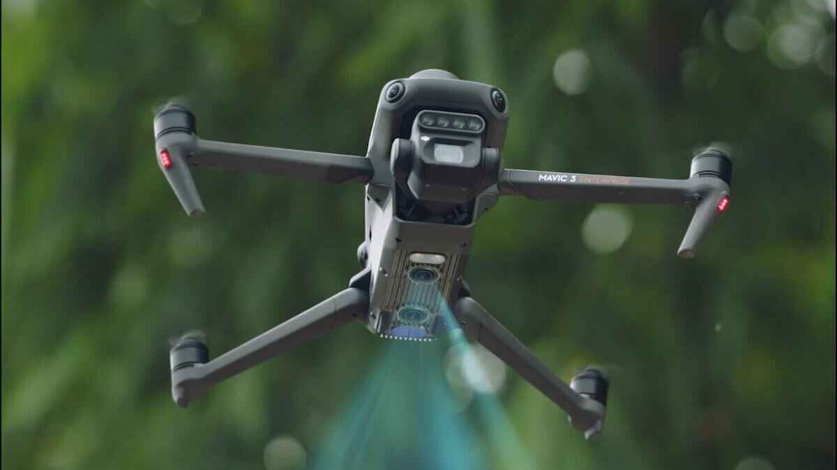 Dron z czterema ramionami i widocznymi kamerami unosi się w powietrzu na tle zieleni. Na jednym z ramion widoczny jest napis \'MAVIC 3\'.