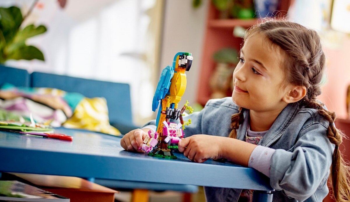 Dziecko siedzi przy stole i bawi się kolorowym modelem papugi z klocków LEGO. W tle widać rozmyte elementy wnętrza mieszkania.