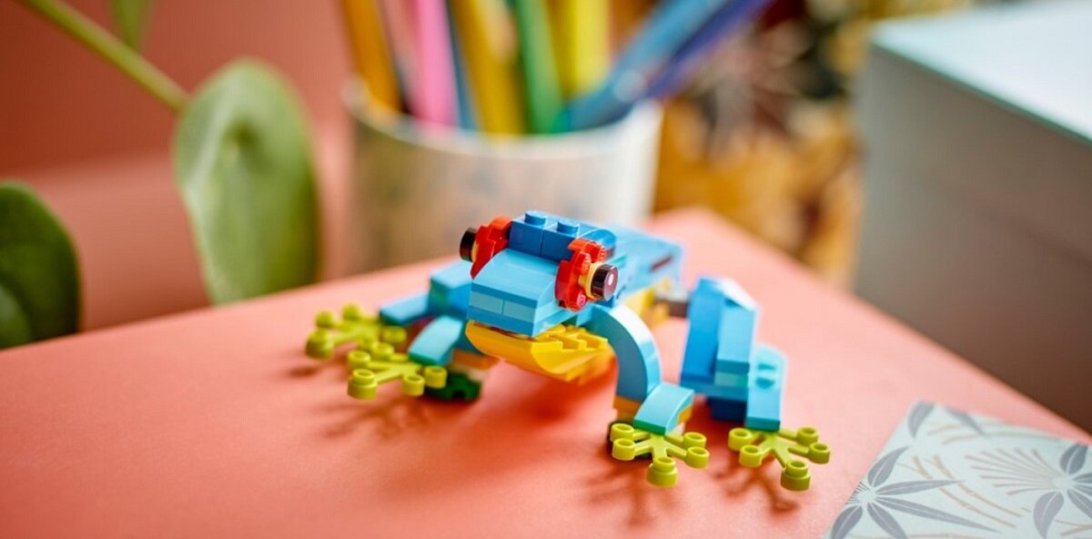 Na obrazie znajduje się kolorowa żaba zbudowana z klocków LEGO, umieszczona na pomarańczowej powierzchni. W tle widoczne są rozmyte elementy, takie jak roślina i przybory do pisania.