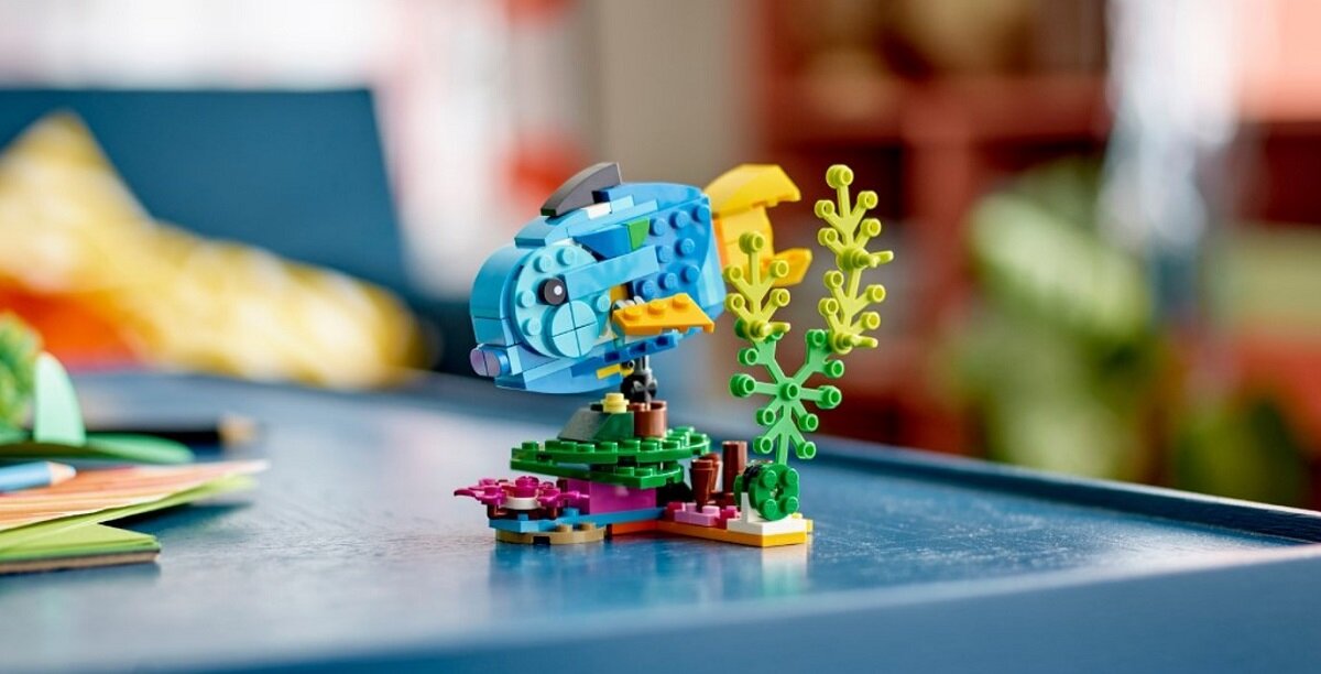 Na stole znajduje się kolorowy model ryby z klocków LEGO, obok której widoczne są elementy przypominające rośliny wodne.
