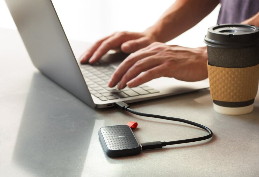 Na stole znajduje się laptop, do którego podłączony jest dysk SANDISK Portable SSD za pomocą kabla USB. Obok laptopa stoi kubek z napojem.