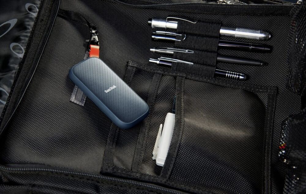 Na obrazie widać przenośny dysk SSD marki SanDisk umieszczony w czarnej torbie z kieszeniami, obok kilku długopisów i innych akcesoriów.