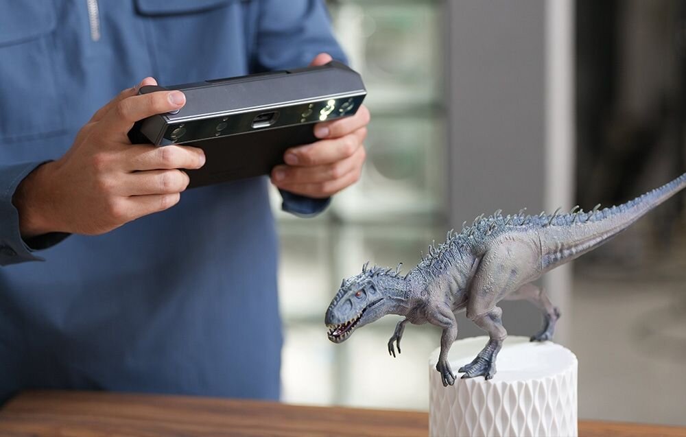 Osoba trzyma urządzenie przypominające skaner 3D, skierowane na model dinozaura stojący na białym postumencie. W tle widoczne są rozmyte elementy wnętrza.