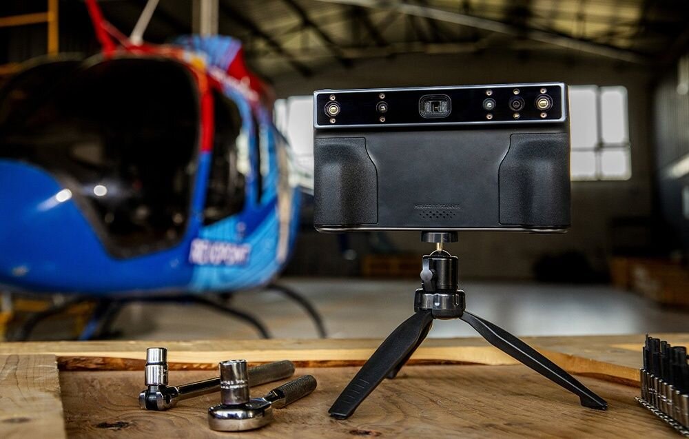 Na drewnianym stole znajduje się czarny skaner 3D na statywie, a w tle widoczny jest niebieski helikopter. Obok skanera leżą narzędzia, takie jak klucze nasadowe.