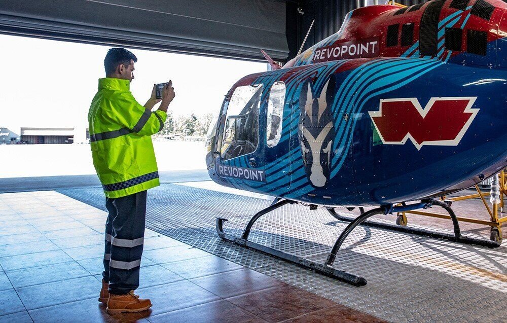 Osoba w jaskrawej kurtce używa urządzenia przed niebieskim helikopterem z logo \'REVOPOINT\'. Helikopter stoi w hangarze z otwartymi drzwiami, a na jego boku widoczny jest biały wzór i czerwone logo w kształcie litery \'W\'.