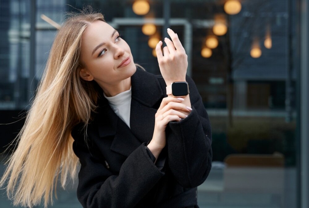 Osoba w ciemnym płaszczu prezentuje smartwatch na nadgarstku. W tle widoczne są rozmyte światła i szklana fasada budynku.