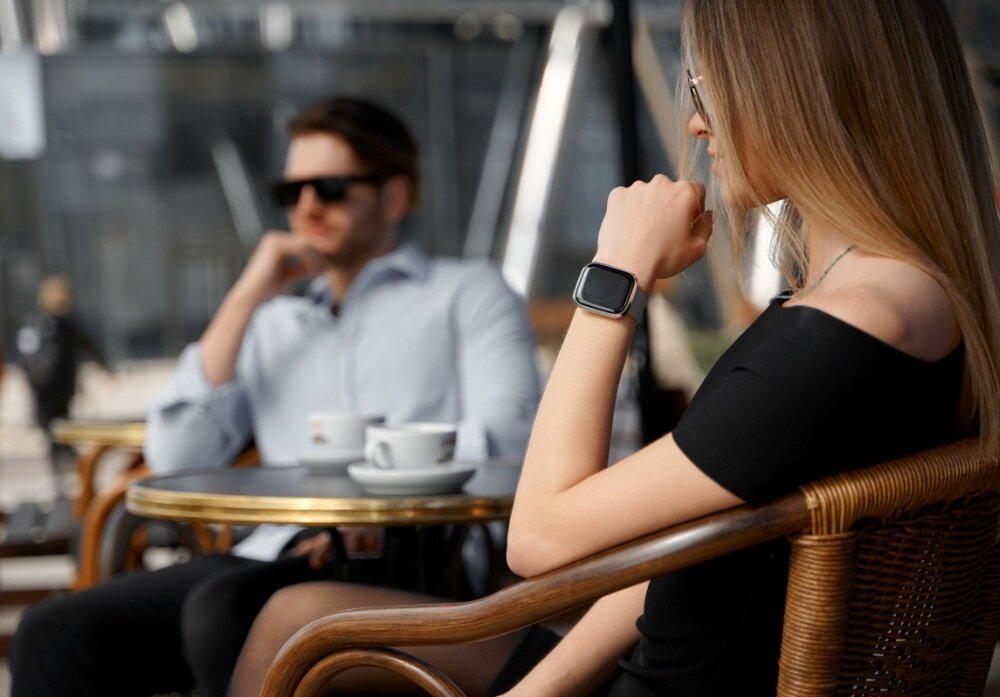 Osoba siedząca przy stoliku w kawiarni nosi srebrny smartwatch na nadgarstku. W tle widać drugą osobę oraz filiżanki z kawą na stoliku.