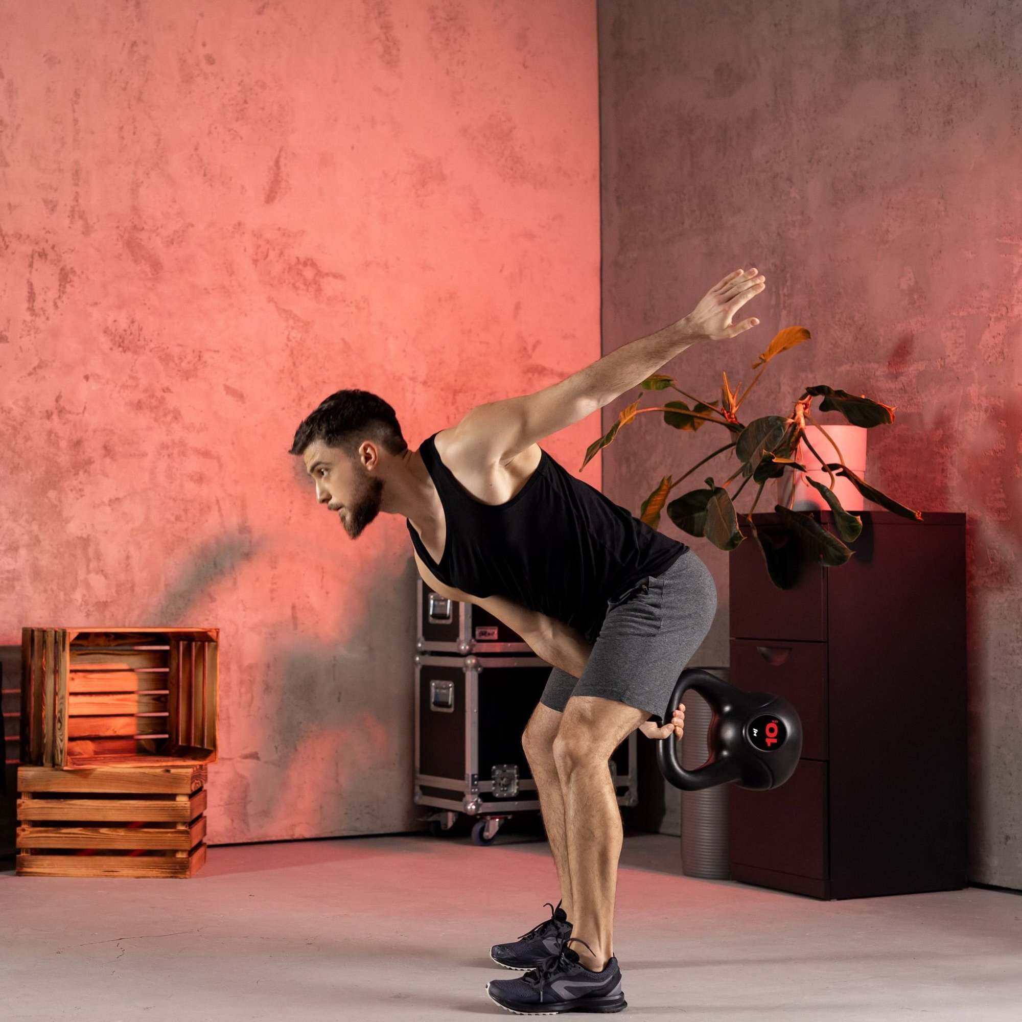 Osoba w czarnym stroju sportowym wykonuje ćwiczenie z czarnym kettlebellem w pomieszczeniu z betonową ścianą i rośliną w tle. Na podłodze stoją drewniane skrzynki i sprzęt muzyczny.