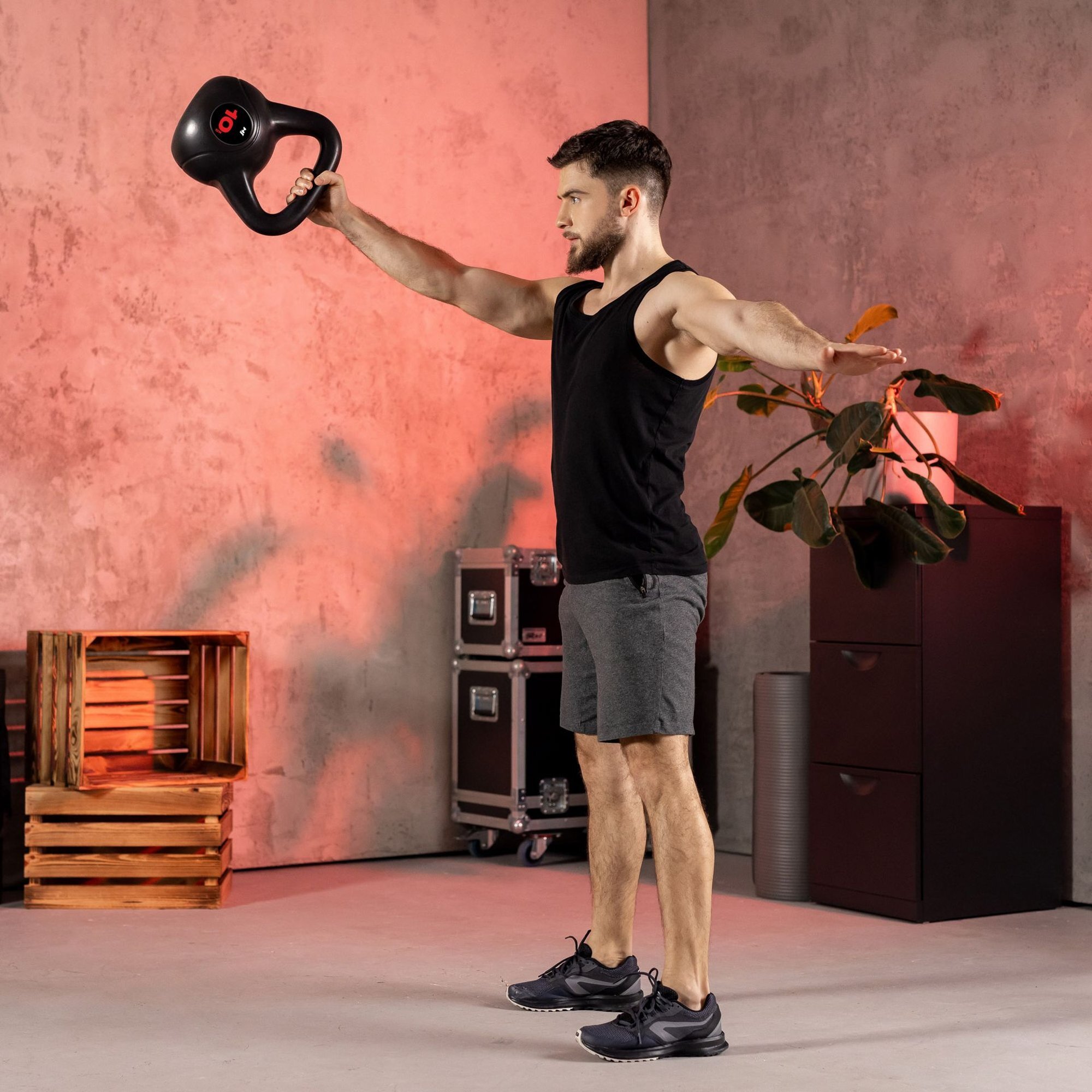 Mężczyzna w czarnej koszulce i szarych szortach trzyma kettlebell w wyciągniętej ręce. W tle widać drewniane skrzynki, metalowe szafki i roślinę doniczkową.