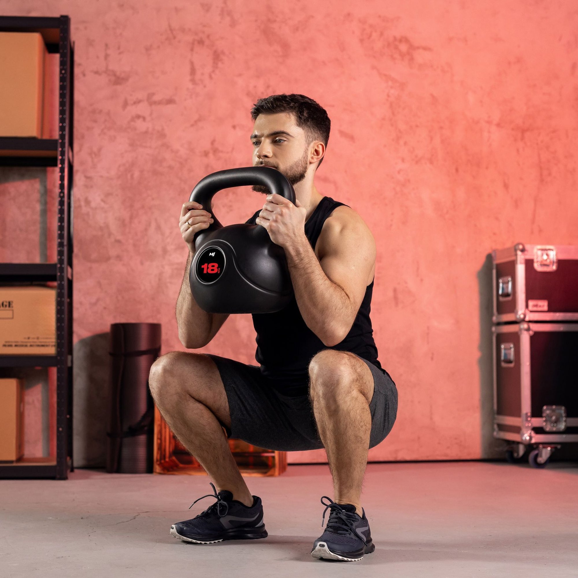 Osoba w pozycji przysiadu trzyma czarny kettlebell z widocznym oznaczeniem \'18\'. W tle widać ścianę i regał z kartonami.