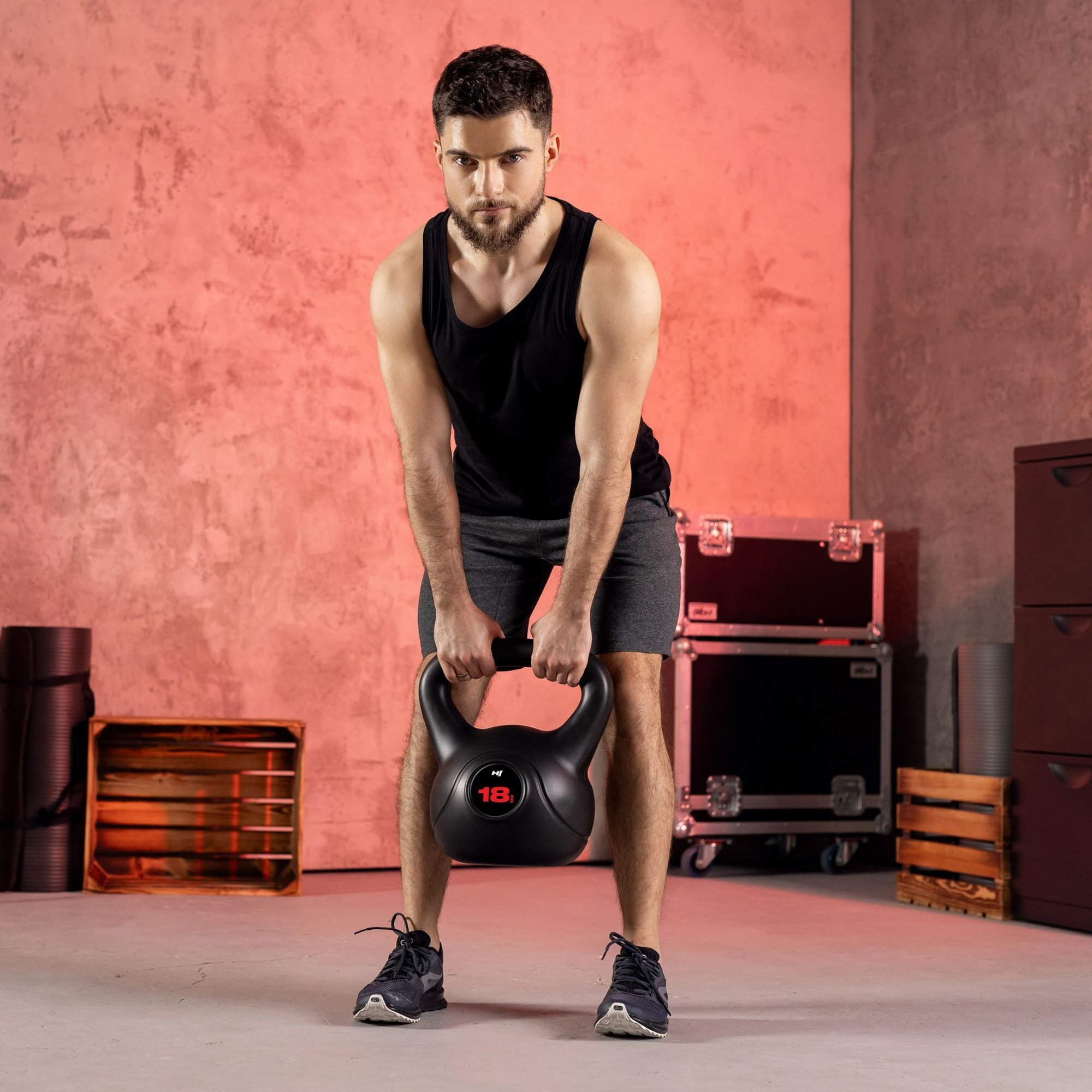 Mężczyzna w czarnej koszulce i szortach trzyma kettlebell winylowy o wadze 18 kg marki HOP SPORT. W tle widoczne są skrzynki i sprzęt sportowy na tle czerwonej ściany.