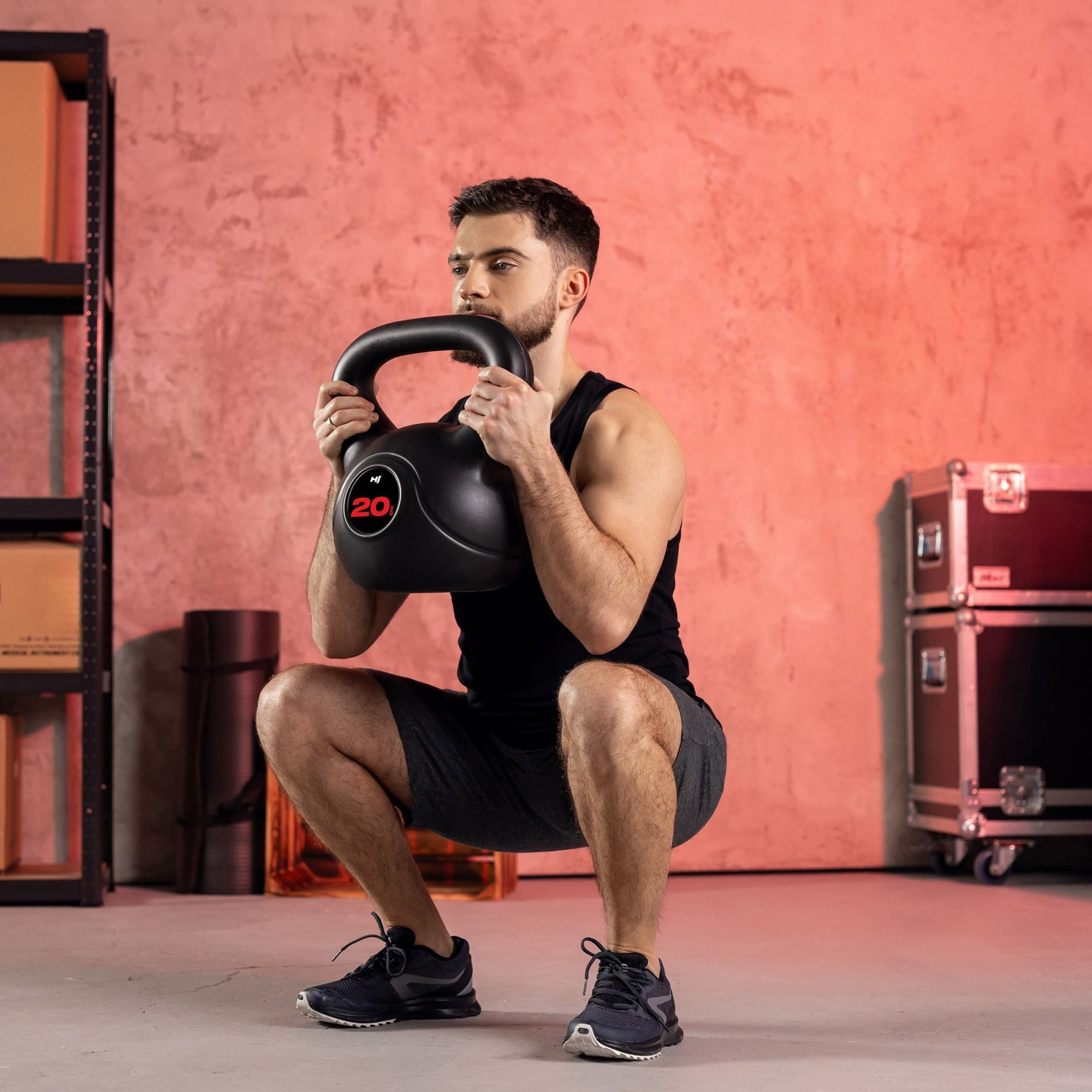 Osoba trzyma czarny kettlebell z czerwonym napisem \'20\' na tle pomieszczenia z różową ścianą i metalowymi regałami.