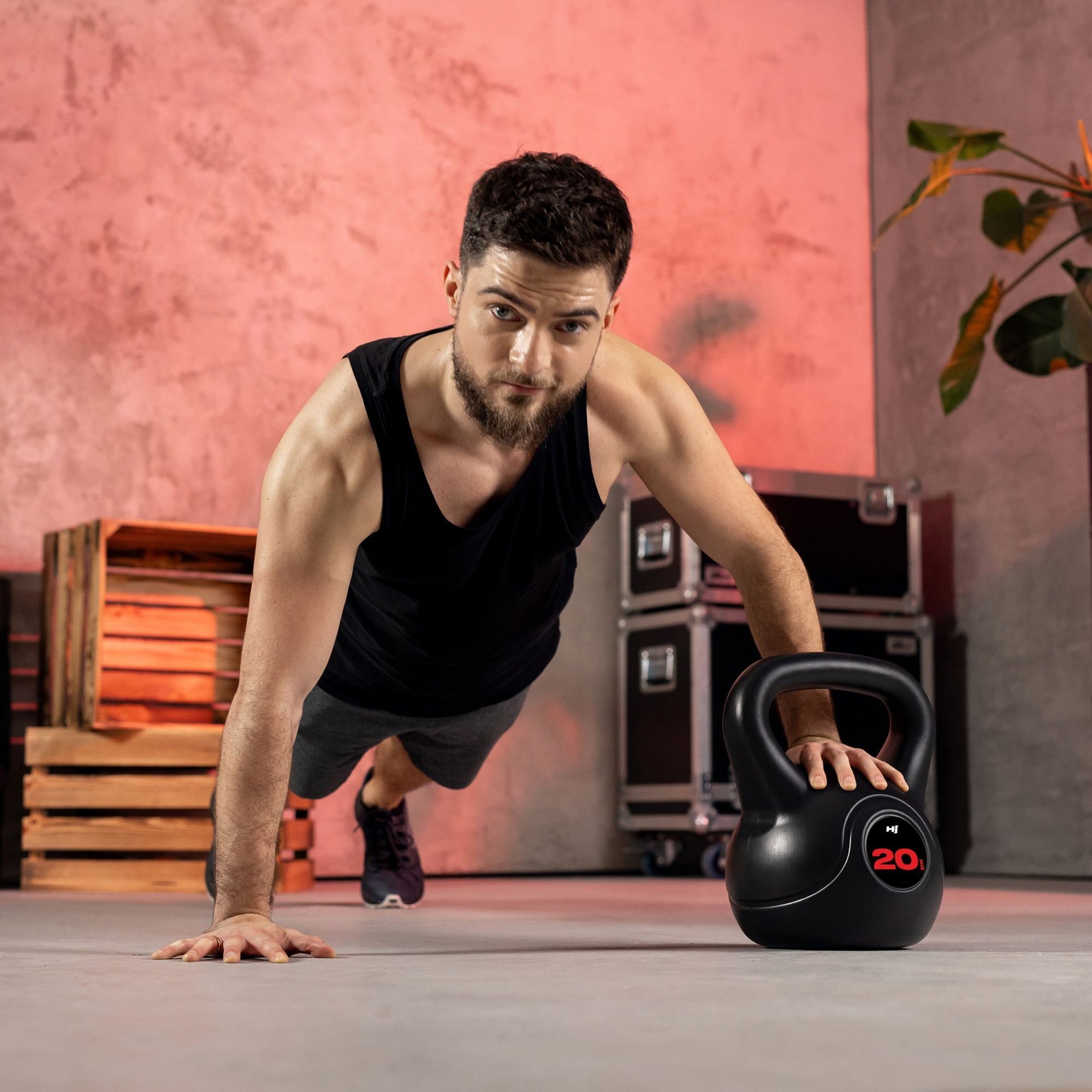 Mężczyzna w czarnej koszulce wykonuje ćwiczenie w pozycji deski, opierając się jedną ręką na czarnym kettlebellu z czerwonym oznaczeniem \'20\'. W tle znajdują się drewniane skrzynki i rośliny na tle ściany w odcieniach szarości i różu.