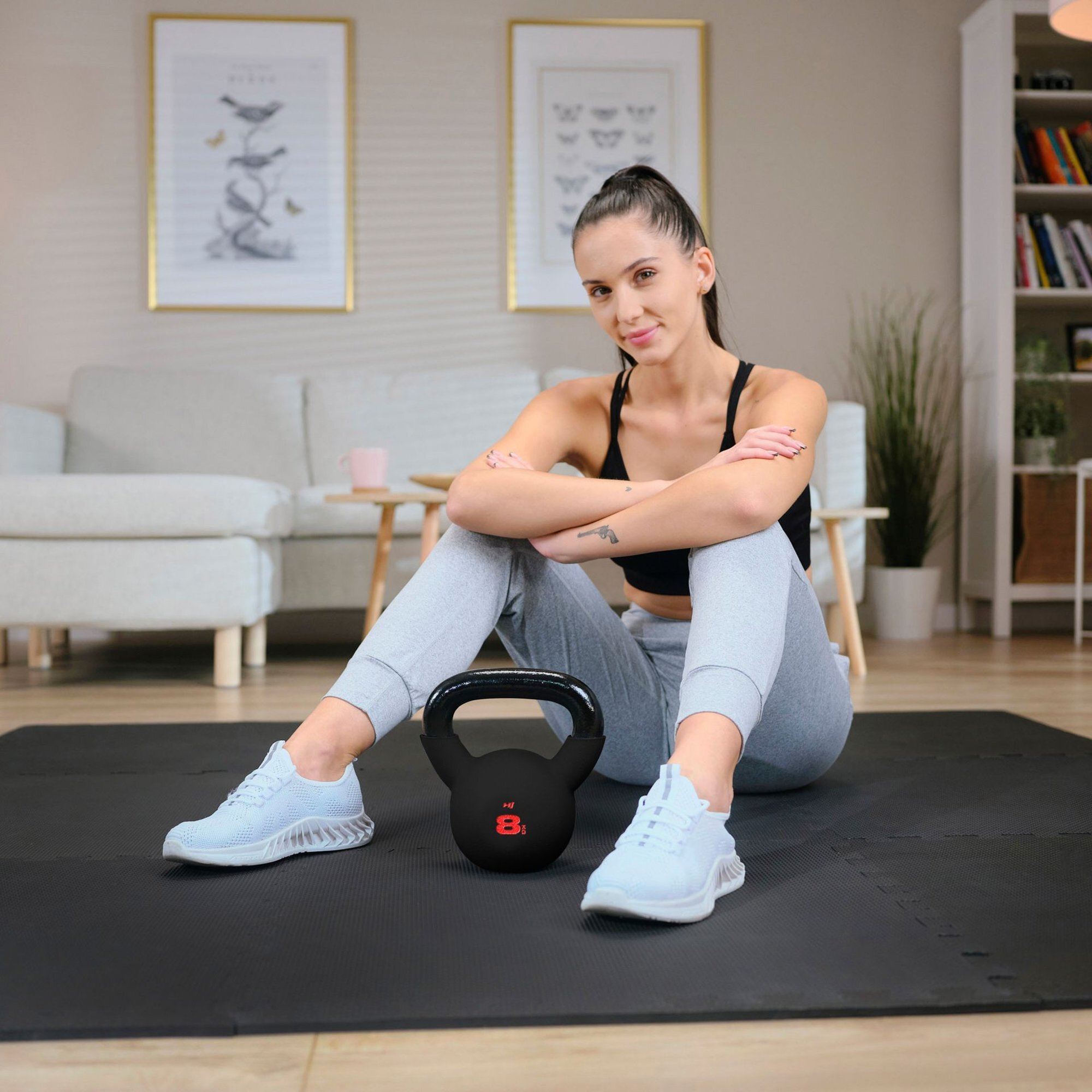 Osoba siedzi na macie do ćwiczeń, obok niej znajduje się czarny odważnik kettlebell z czerwonym napisem \'8 kg\'. W tle widoczny jest jasny salon z sofą, stolikiem i rośliną.