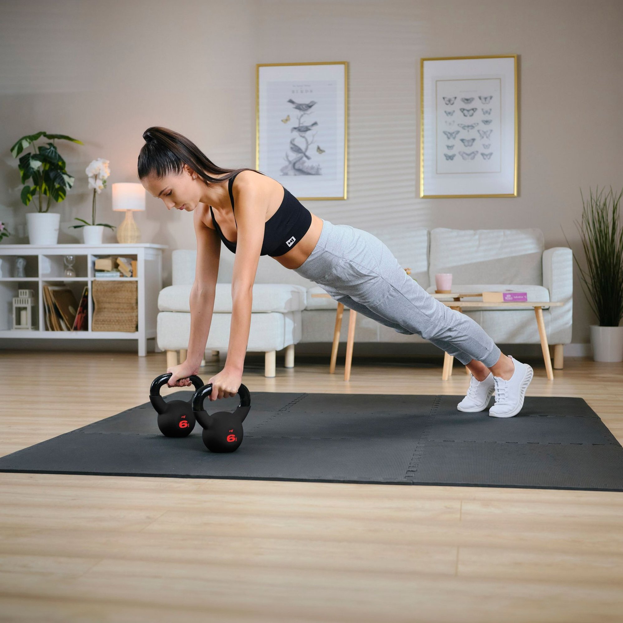 Osoba wykonuje ćwiczenie w pozycji deski, opierając się na dwóch czarnych odważnikach kettlebell z czerwonym oznaczeniem \'6\'. W tle znajduje się jasne wnętrze z sofą, stolikiem i roślinami.