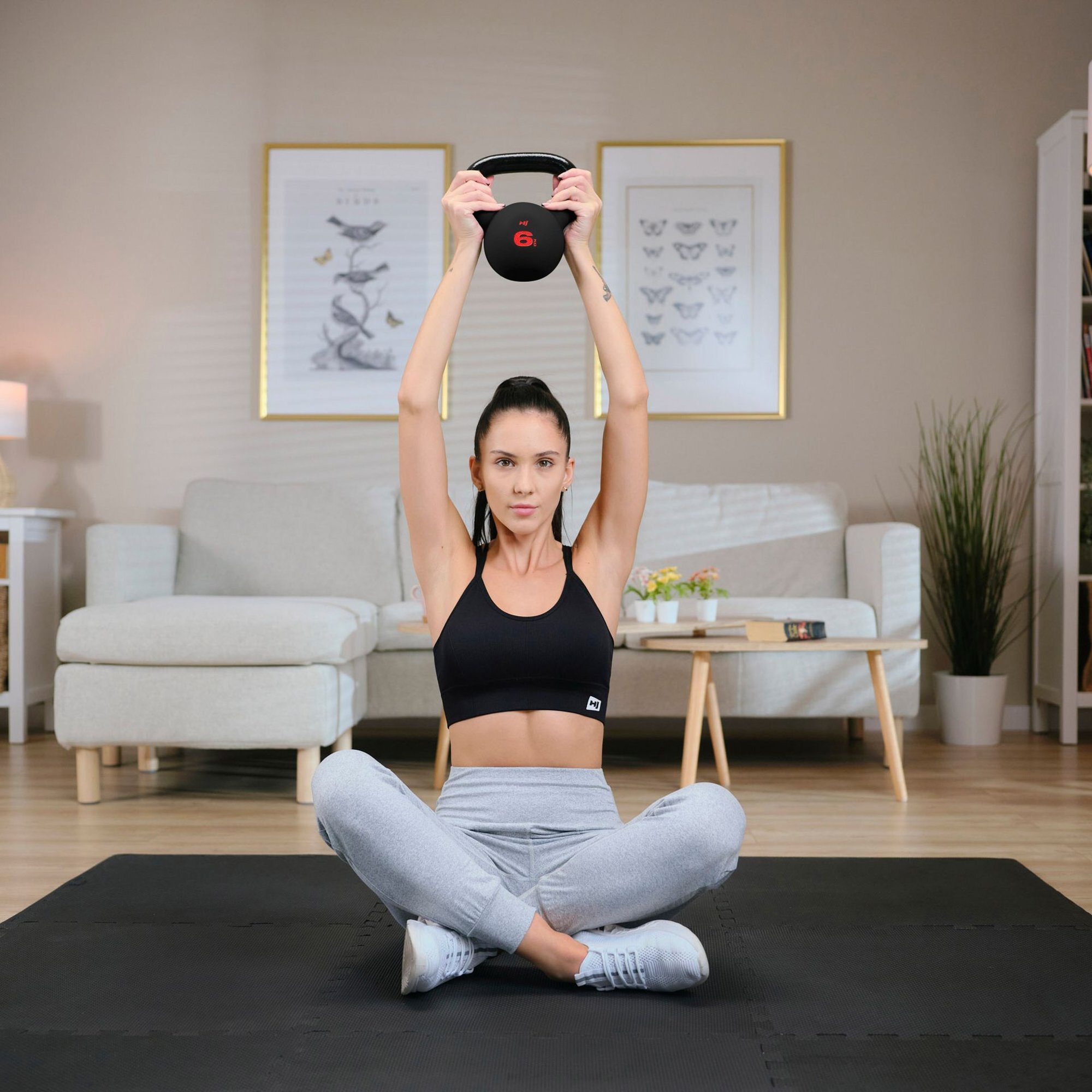 Osoba siedzi na macie w salonie, trzymając nad głową czarny odważnik kettlebell z czerwonym napisem \'6\'. W tle widoczne są kanapa, stolik z książką i rośliną oraz dwa obrazy na ścianie.