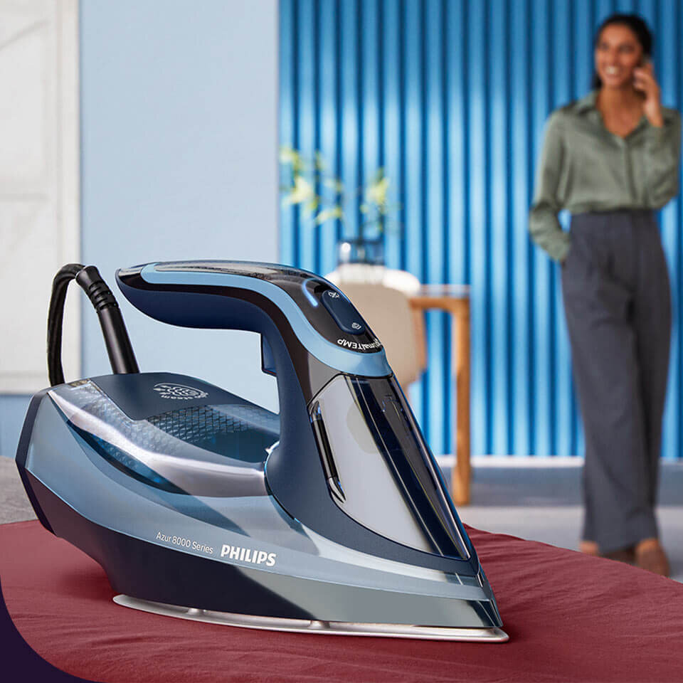 Na pierwszym planie znajduje się żelazko PHILIPS Azur 8000 Series DST8020/20 w kolorze niebieskim. W tle widoczna jest osoba stojąca w pomieszczeniu z niebieskimi ścianami.