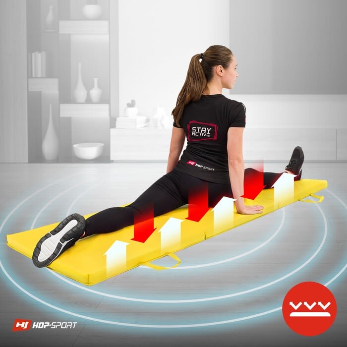 Osoba siedzi na żółtym, składanym materacu gimnastycznym marki HOP-SPORT, wykonując ćwiczenie rozciągające. Na materacu widoczne są czerwone i białe strzałki skierowane w górę, a w tle znajduje się wnętrze pokoju z półkami i dekoracjami.