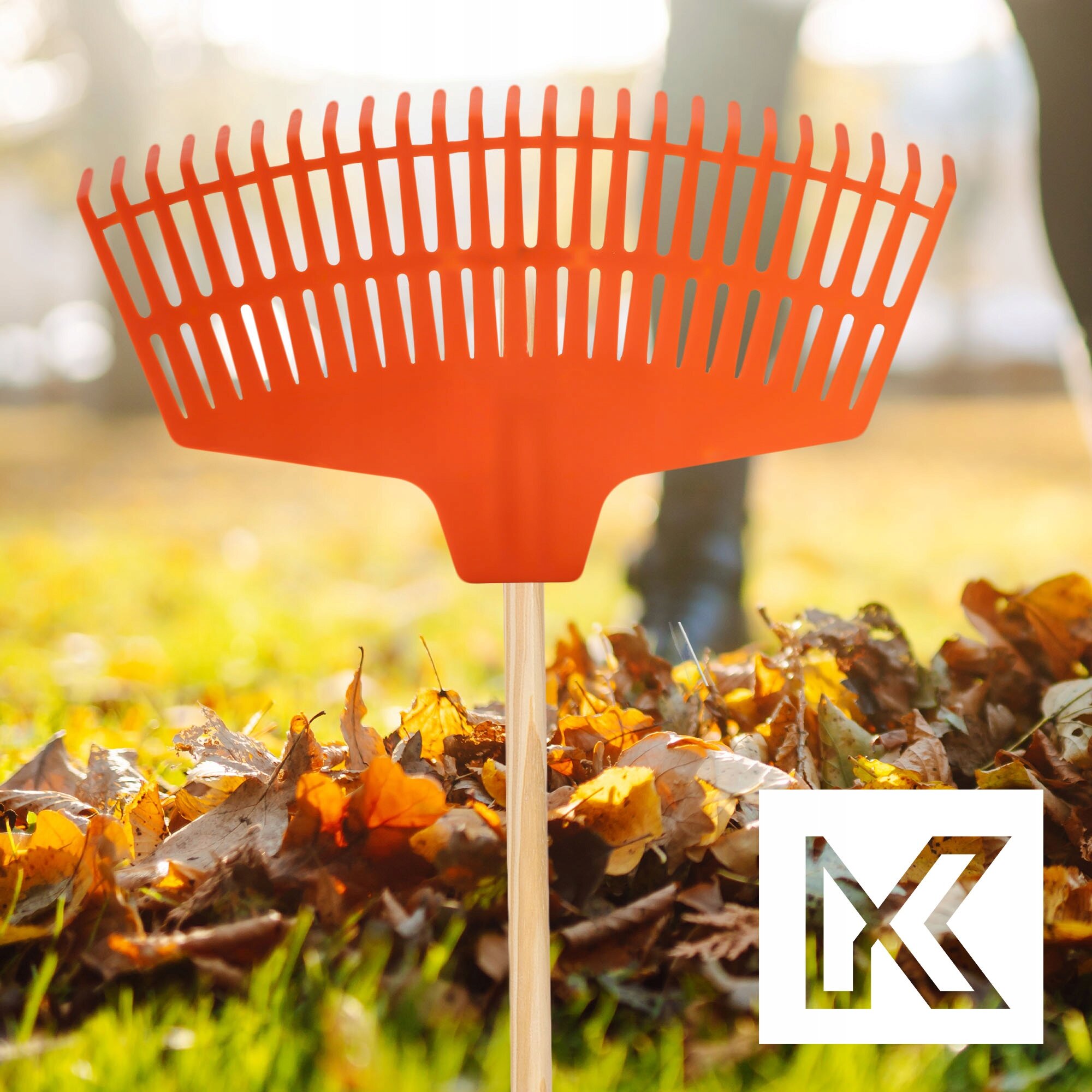 Pomarańczowe plastikowe grabie ogrodowe do liści bez trzonka na tle jesiennych liści. W prawym dolnym rogu widoczne jest logo w kształcie litery K.