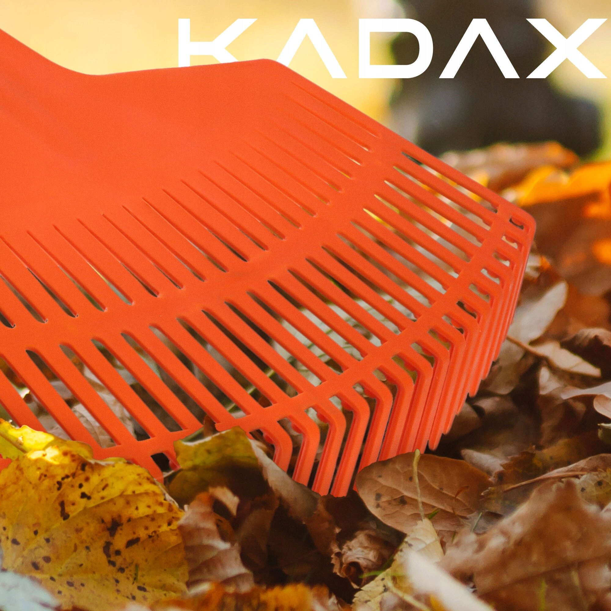 Pomarańczowe plastikowe grabie ogrodowe do liści leżą na stosie suchych liści. Widoczne jest logo \'KADAX\'.
