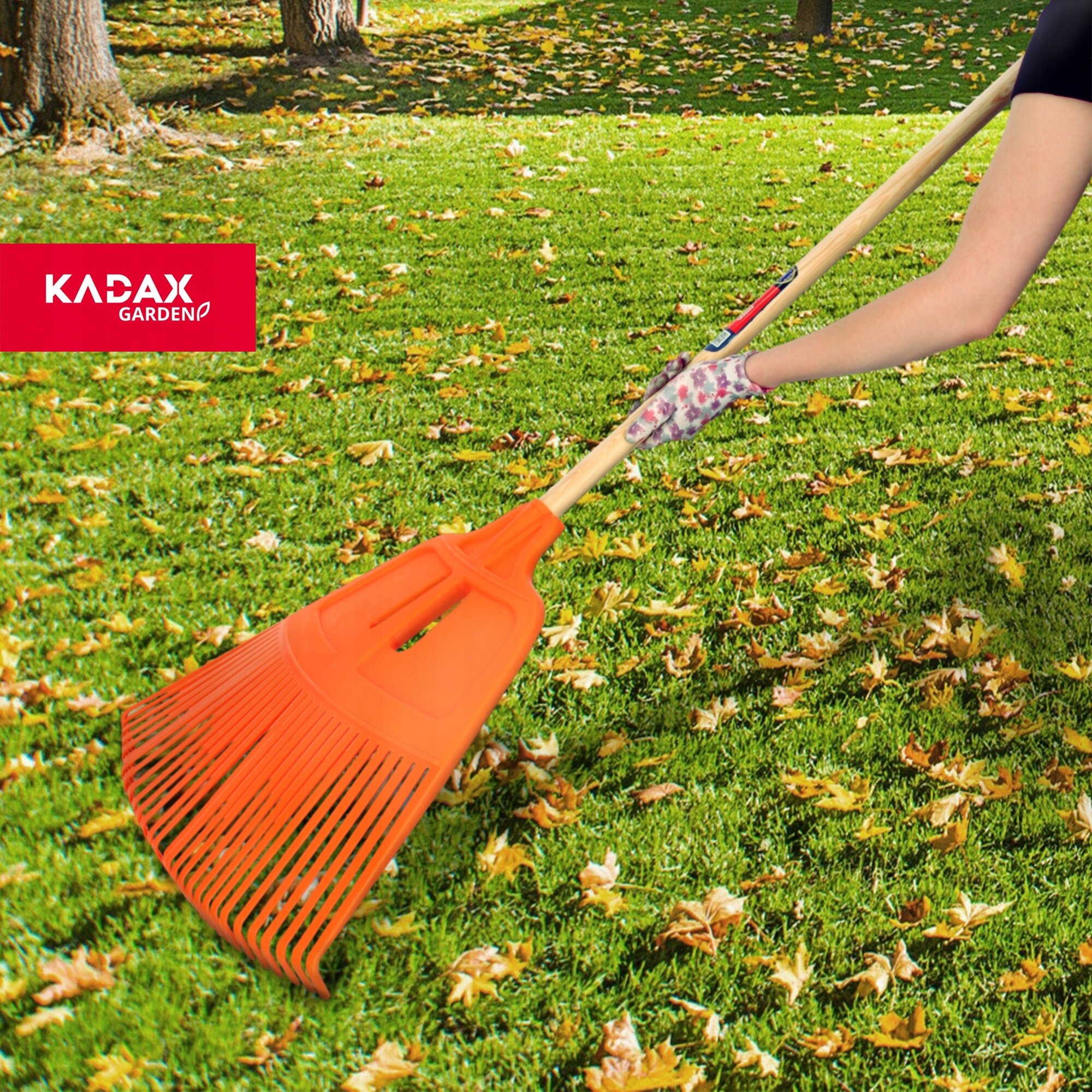 Osoba trzyma pomarańczowe grabie wachlarzowe na tle trawnika pokrytego liśćmi. Widoczne jest logo \'KADAX GARDEN\'.