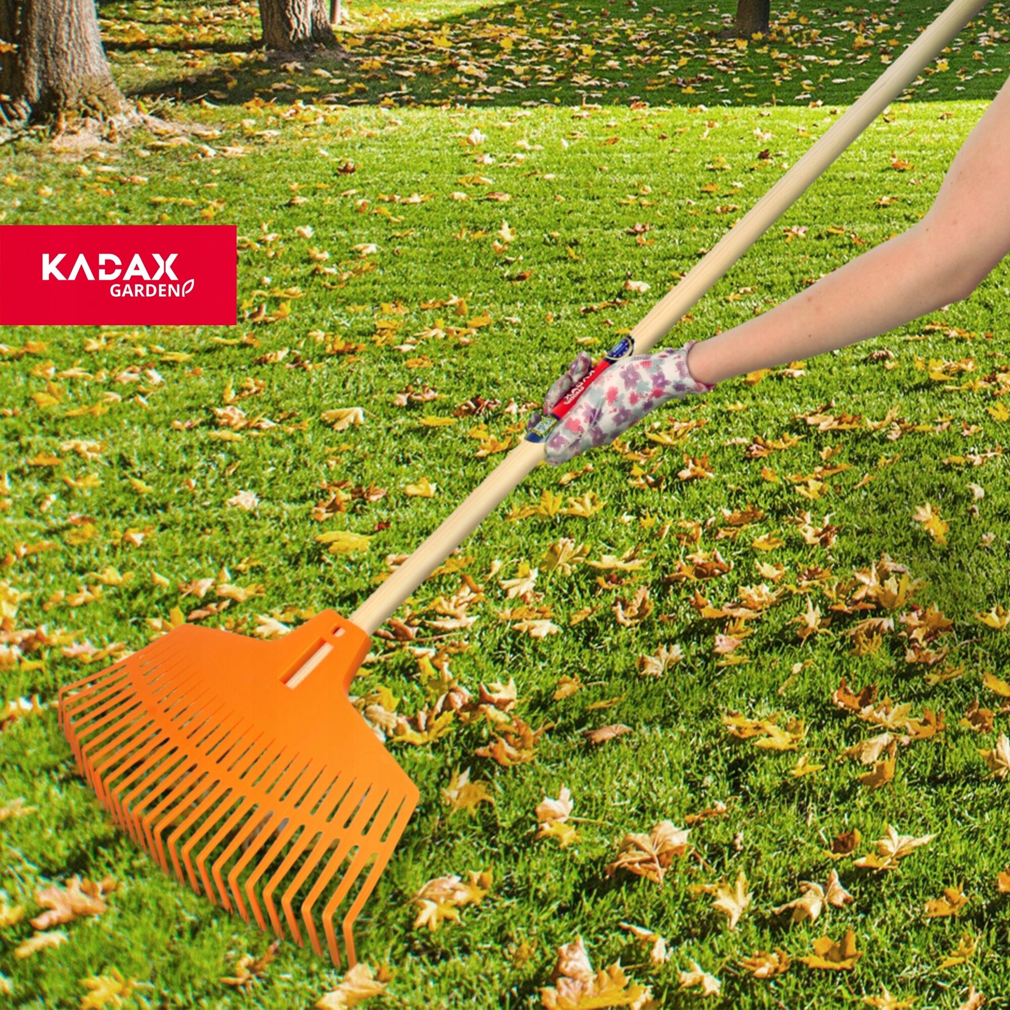 Osoba trzyma grabie z drewnianym trzonkiem i pomarańczową głowicą na trawniku pokrytym liśćmi. Widoczne logo: KADAX GARDEN.