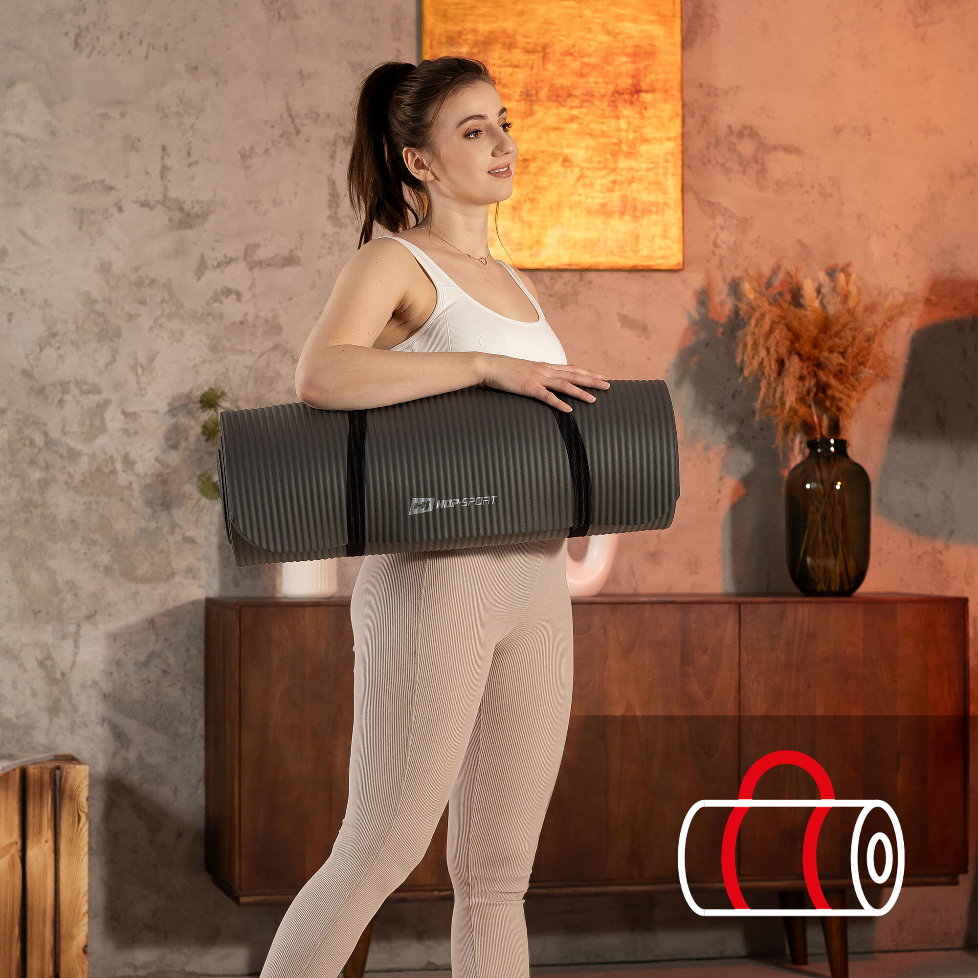 Osoba trzyma szarą matę fitness z logo \'HOP SPORT\' na tle wnętrza z drewnianą komodą i dekoracjami. W prawym dolnym rogu znajduje się ikona przedstawiająca zwiniętą matę z uchwytem.