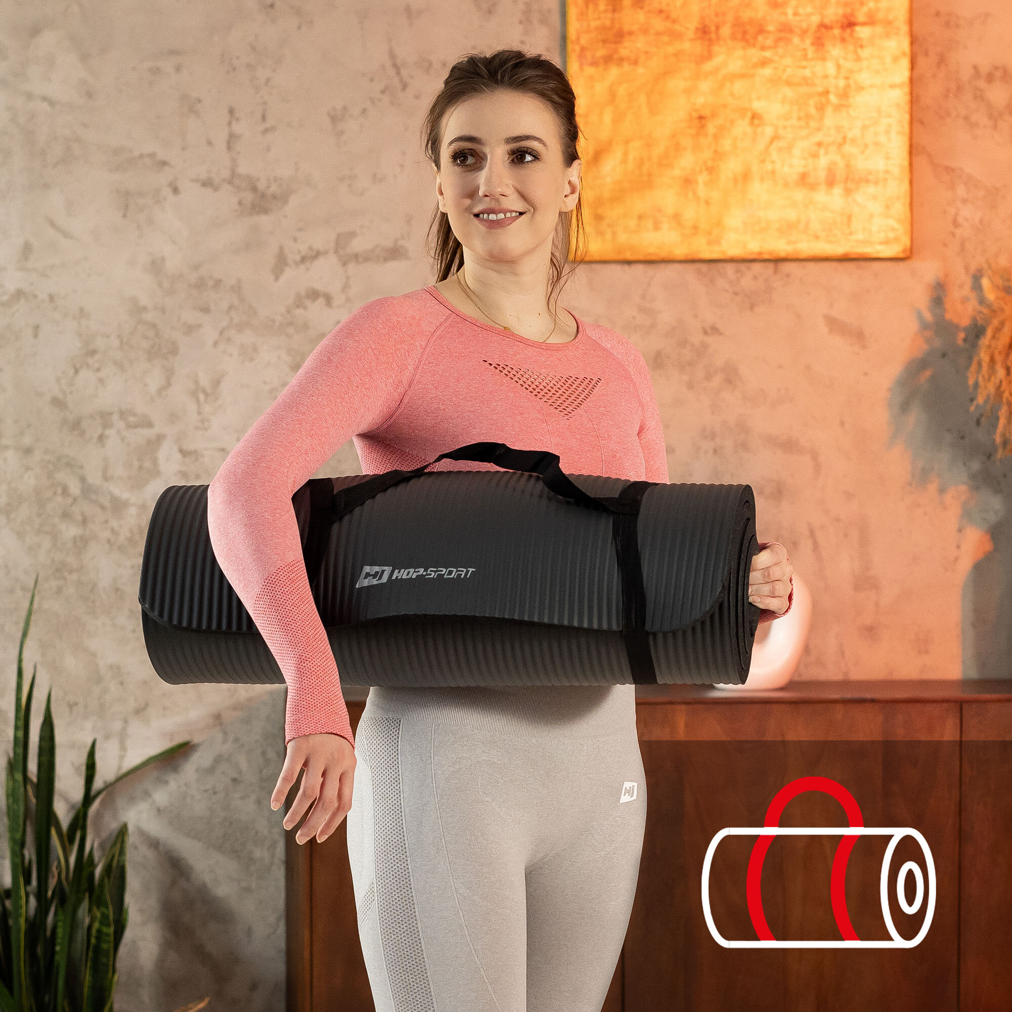 Osoba trzyma czarną matę do ćwiczeń z logo \'HOP-SPORT\'. W tle widoczna jest ściana i roślina, a w prawym dolnym rogu znajduje się ikona przedstawiająca zwiniętą matę z uchwytem.