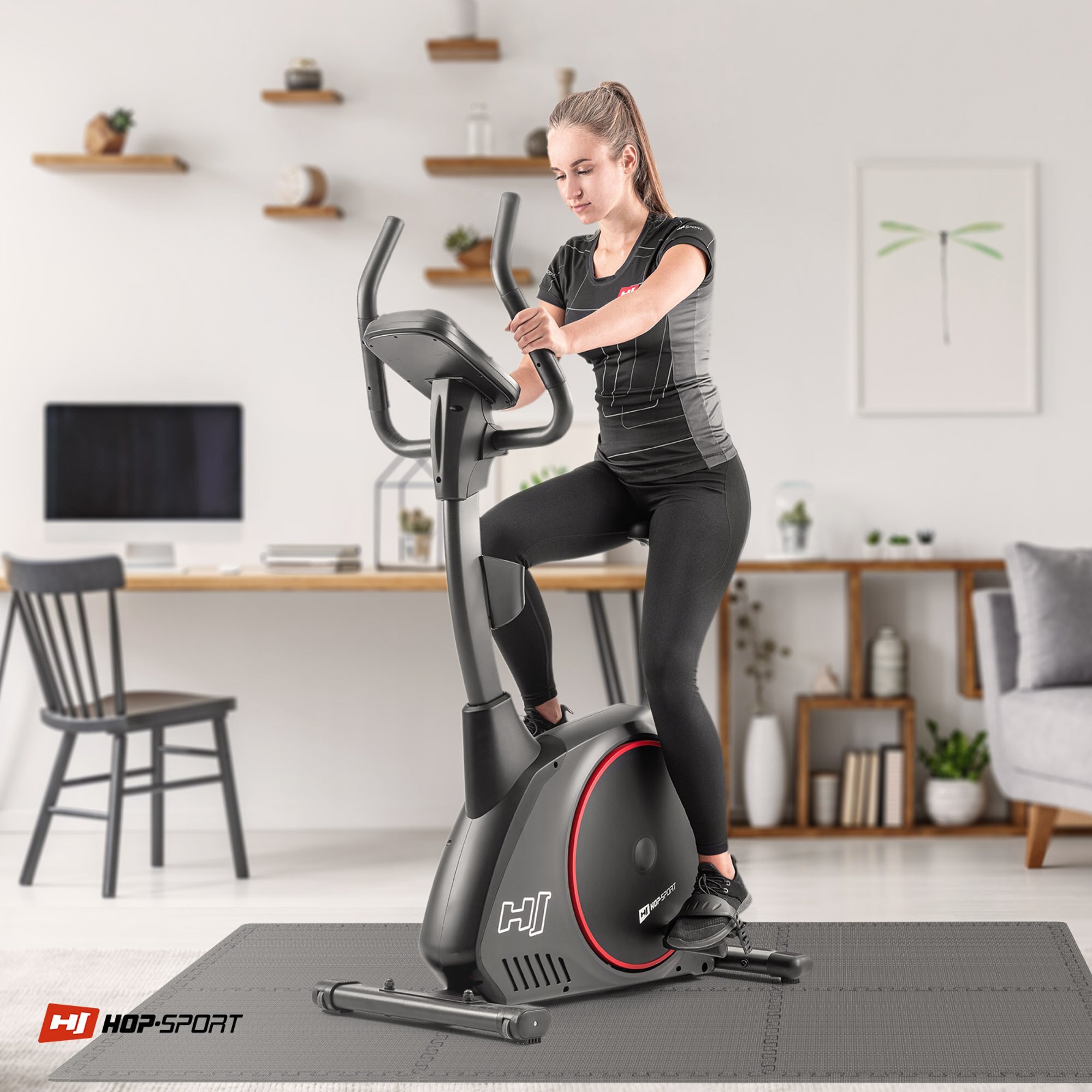 Osoba ćwiczy na rowerze stacjonarnym w jasnym pomieszczeniu z półkami i biurkiem w tle. Na podłodze znajduje się szara mata puzzle, a w rogu widoczne jest logo \'HOP-SPORT\'.