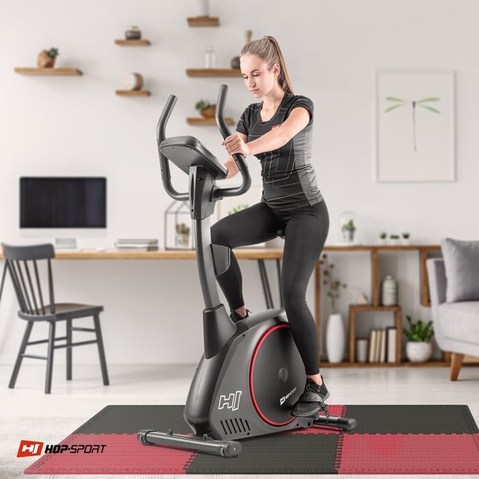 Osoba ćwiczy na rowerze stacjonarnym umieszczonym na macie puzzle EVA w kolorach czarnym i czerwonym. W tle znajduje się biurko z krzesłem oraz półki z roślinami i dekoracjami.