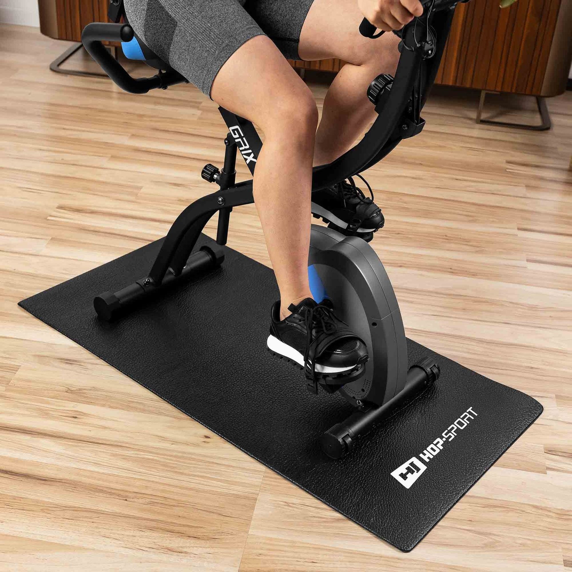 Osoba ćwiczy na rowerze stacjonarnym ustawionym na czarnej macie z logo \'HOP-SPORT\'. Mata znajduje się na drewnianej podłodze.