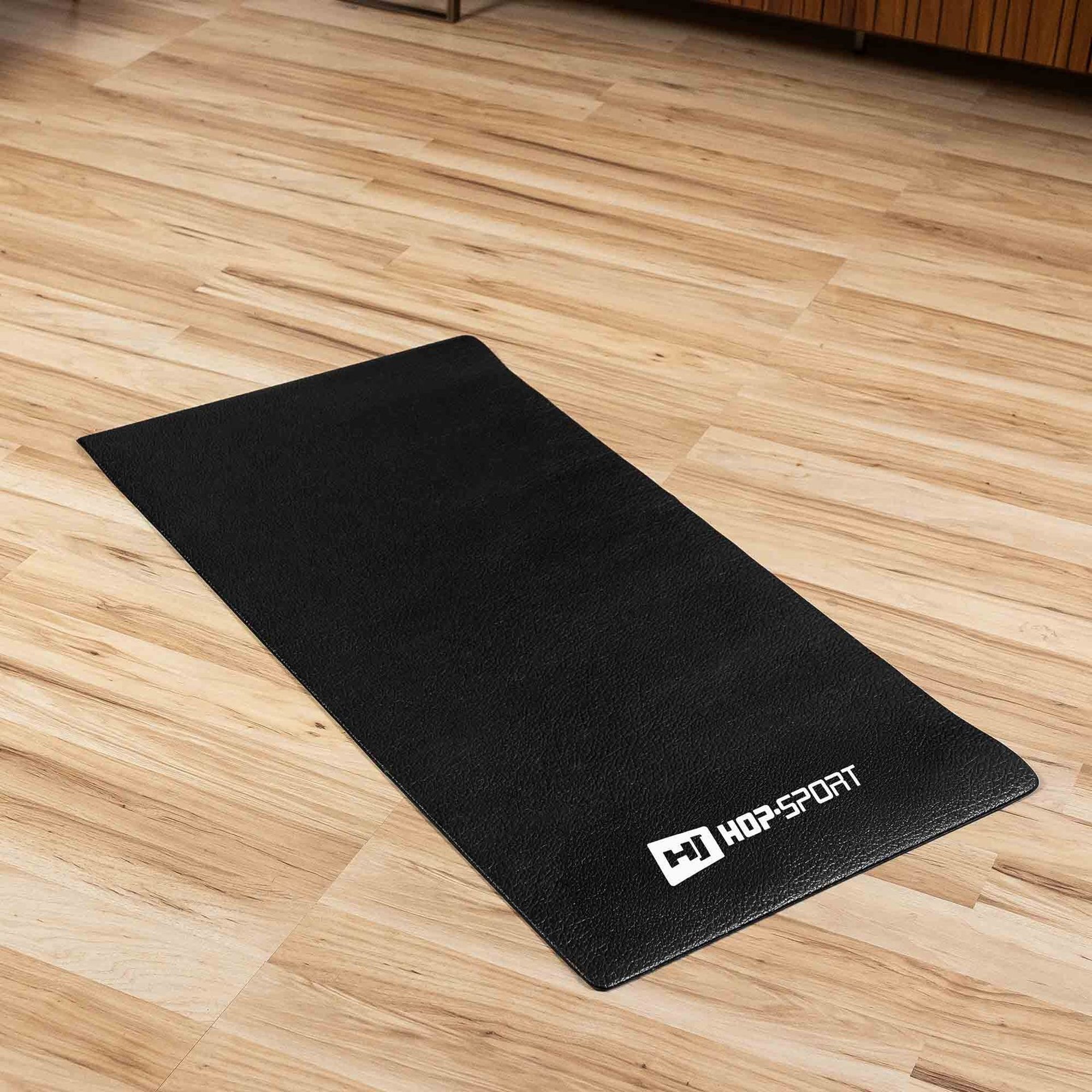 Czarna mata do ćwiczeń leży na drewnianej podłodze. Na macie widoczne jest białe logo z napisem \'HOP-SPORT\'.