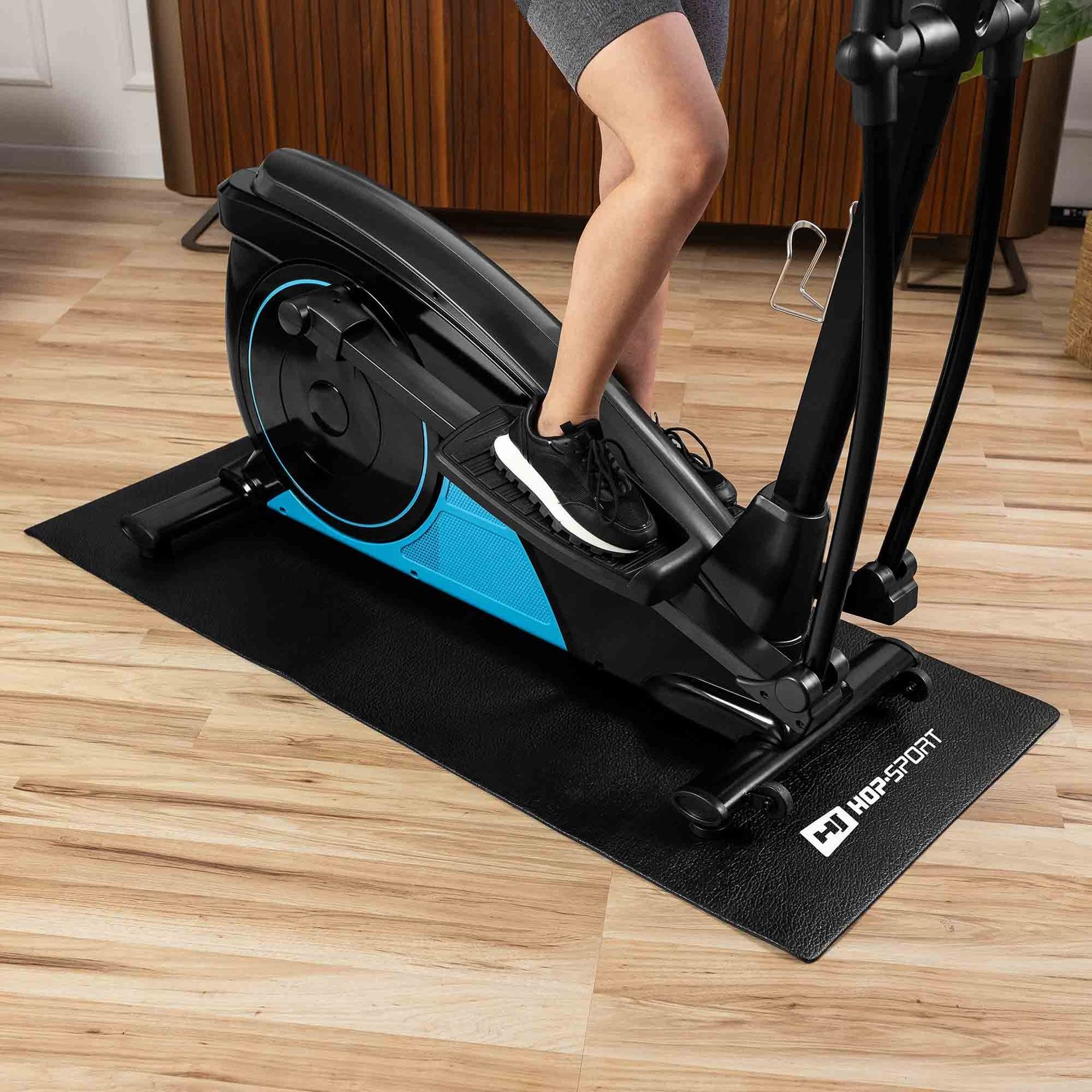 Osoba ćwiczy na czarnym sprzęcie fitness ustawionym na czarnej macie z logo \'HOP-SPORT\'. Podłoga jest drewniana, a w tle widać drewnianą szafkę.