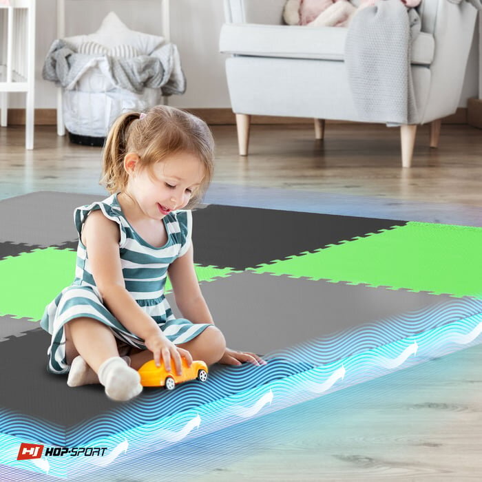 Dziewczynka bawi się żółtym samochodzikiem na macie puzzle w kolorach czarnym, szarym i zielonym. W rogu widoczne jest logo \'HOP-SPORT\'.