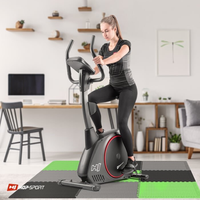 Osoba ćwiczy na rowerze stacjonarnym, stojącym na macie puzzle w kolorach czarnym, szarym i zielonym. W tle widać biurko z krzesłem oraz półki z dekoracjami, a na macie znajduje się logo \'HOP-SPORT\'.