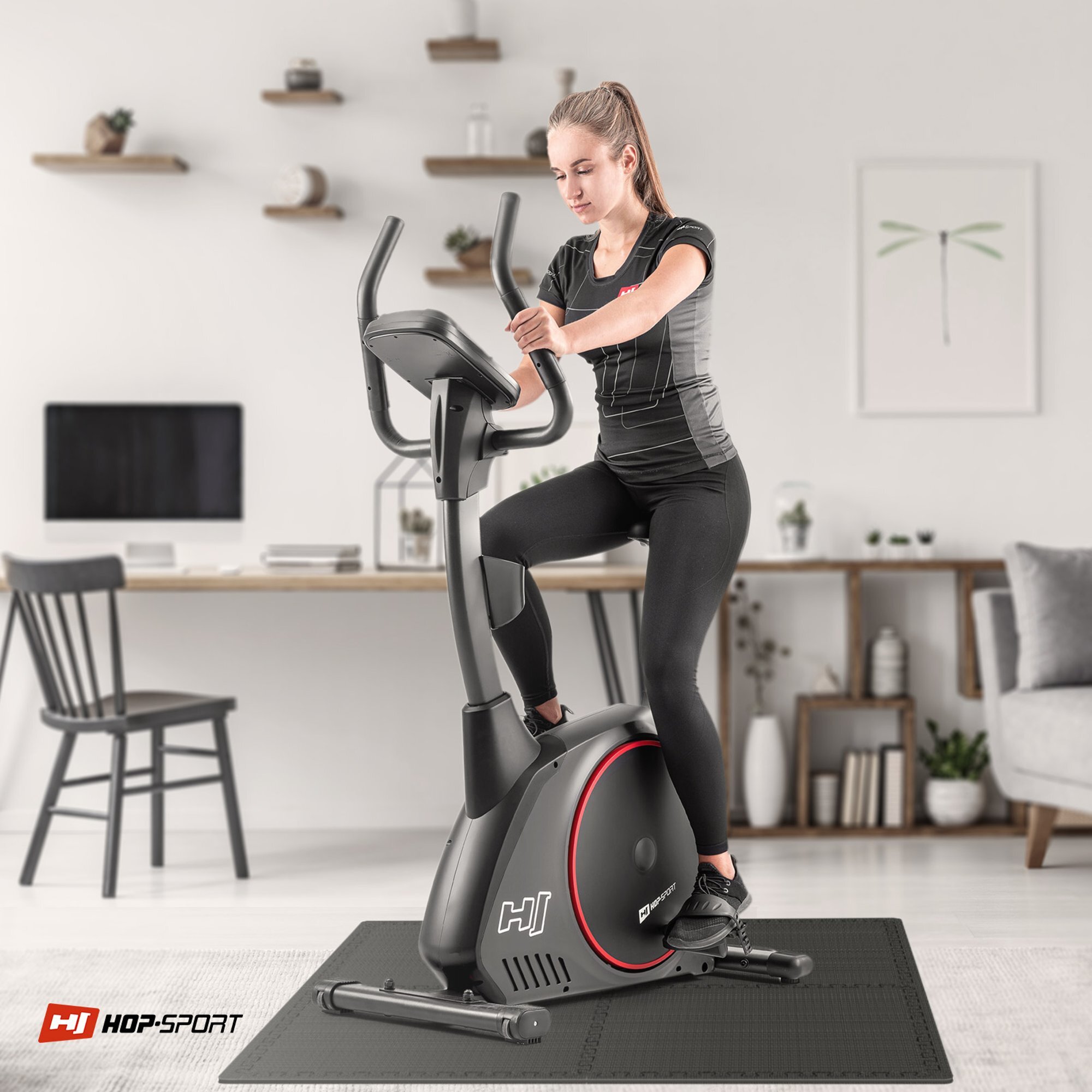 Osoba ćwiczy na rowerze stacjonarnym umieszczonym na czarnej macie puzzle EVA. W tle znajduje się biurko z komputerem i dekoracje ścienne, a na macie widoczne jest logo \'HOP-SPORT\'.