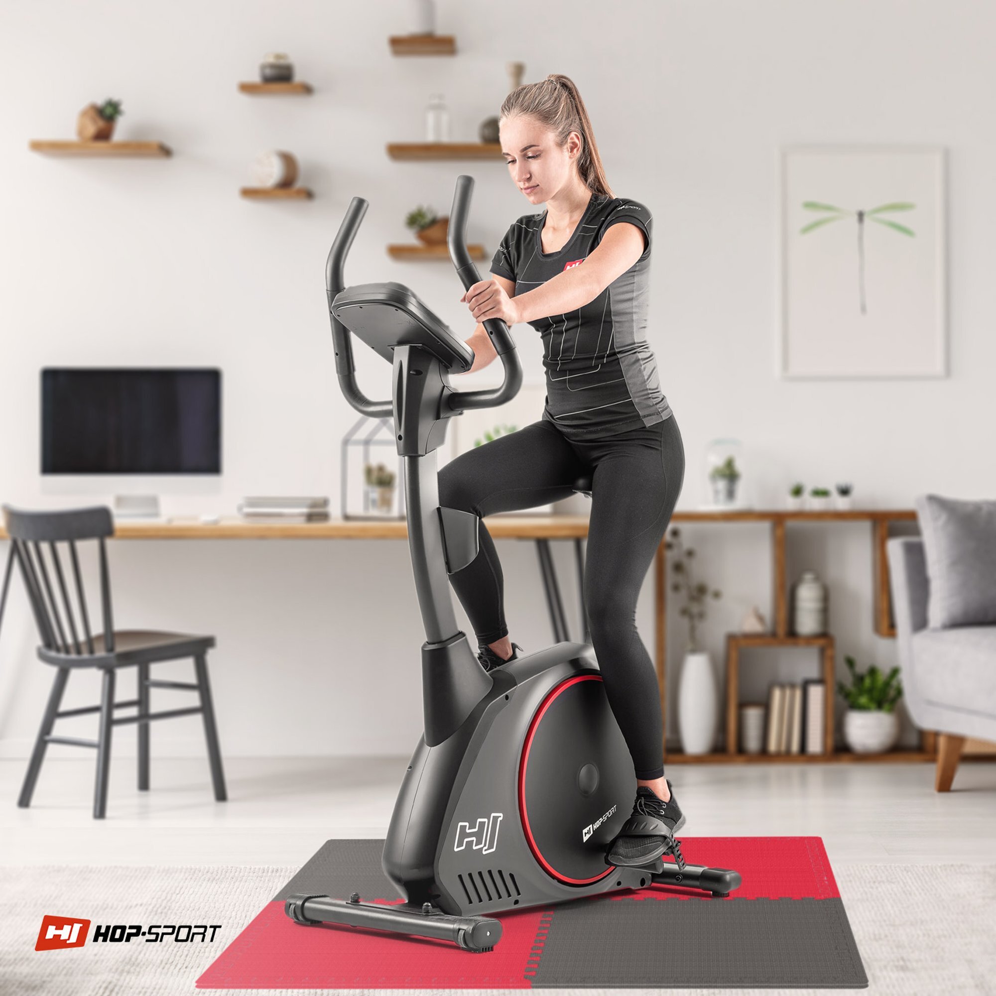 Osoba ćwiczy na rowerze stacjonarnym umieszczonym na macie puzzle EVA w kolorach czarnym i czerwonym. W tle znajduje się biurko z krzesłem oraz półki z dekoracjami, a na macie widoczne jest logo HOP-SPORT.