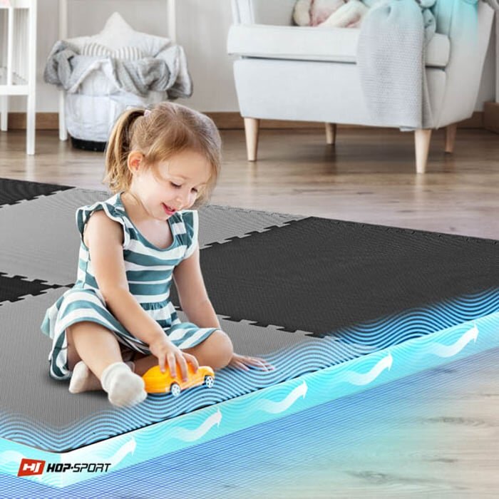 Dziecko siedzi na macie puzzle w kolorach czarnym i szarym, bawiąc się żółtym samochodzikiem. W lewym dolnym rogu widoczne jest logo \'HOP-SPORT\'.