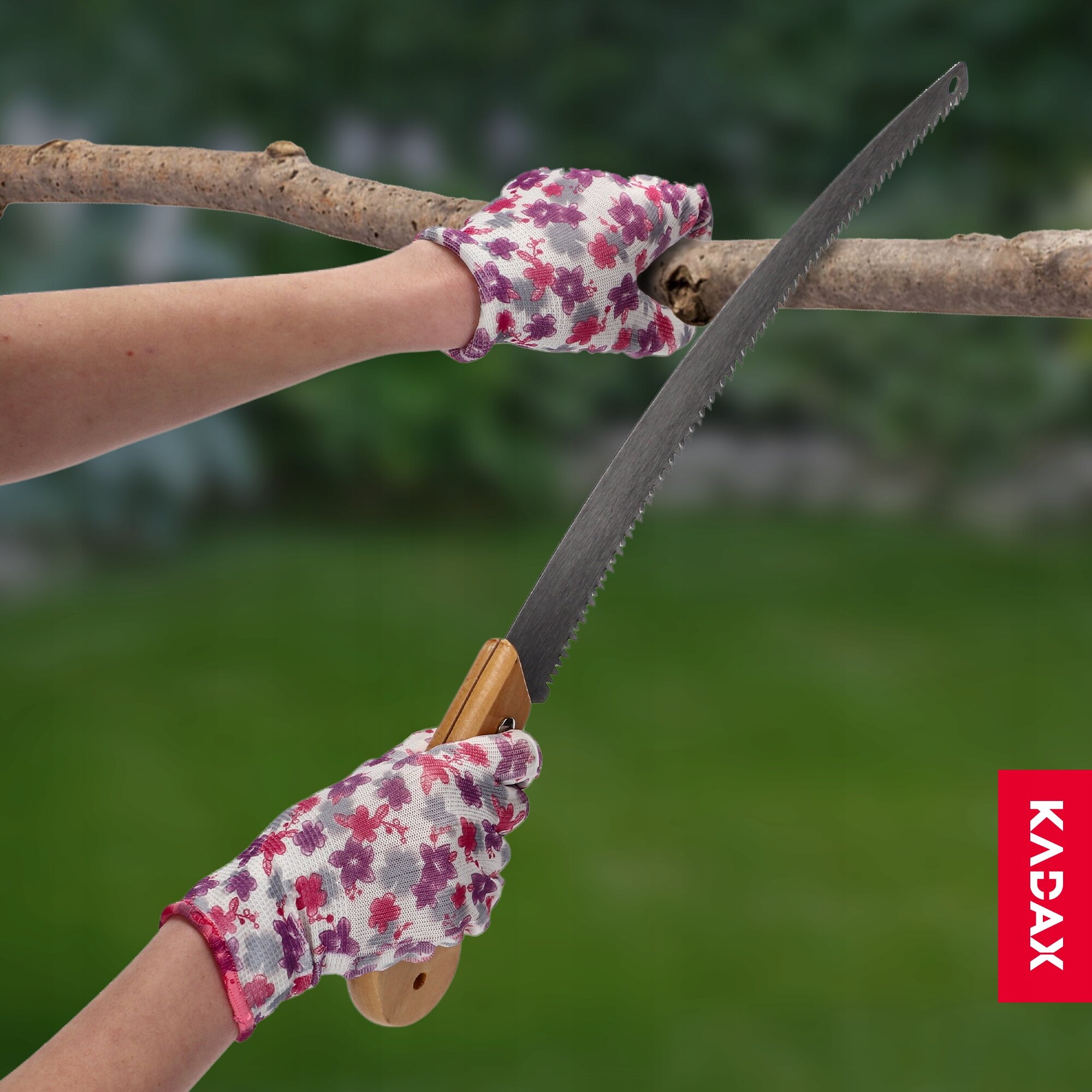 Osoba w rękawiczkach z motywem kwiatowym trzyma piłę ręczną, przecinając gałąź. W prawym dolnym rogu widoczne jest logo \'KADAX\'.