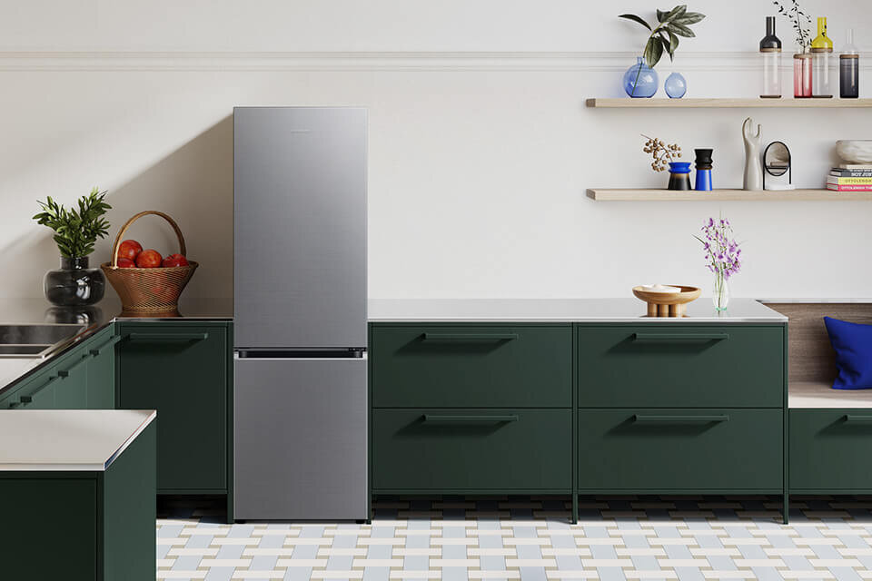Lodówka Samsung Combi AI RB34C603CS9 dostępna w Media Expert W kuchni znajduje się lodówka ze szczotkowanej stali nierdzewnej, otoczona zielonymi szafkami. Na blacie i półkach widoczne są różne przedmioty, w tym kosz z owocami i wazony.
