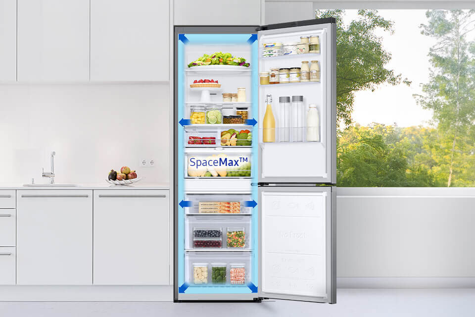 lodówka z dolnym zamrażalnikiem rb34c603cs9 z technologią spacemax Otwarta lodówka z widocznymi półkami wypełnionymi różnymi produktami spożywczymi, na drzwiach napis \'SpaceMax™\'. W tle znajduje się jasna kuchnia z blatem i zlewem oraz widok na zieleń za oknem.