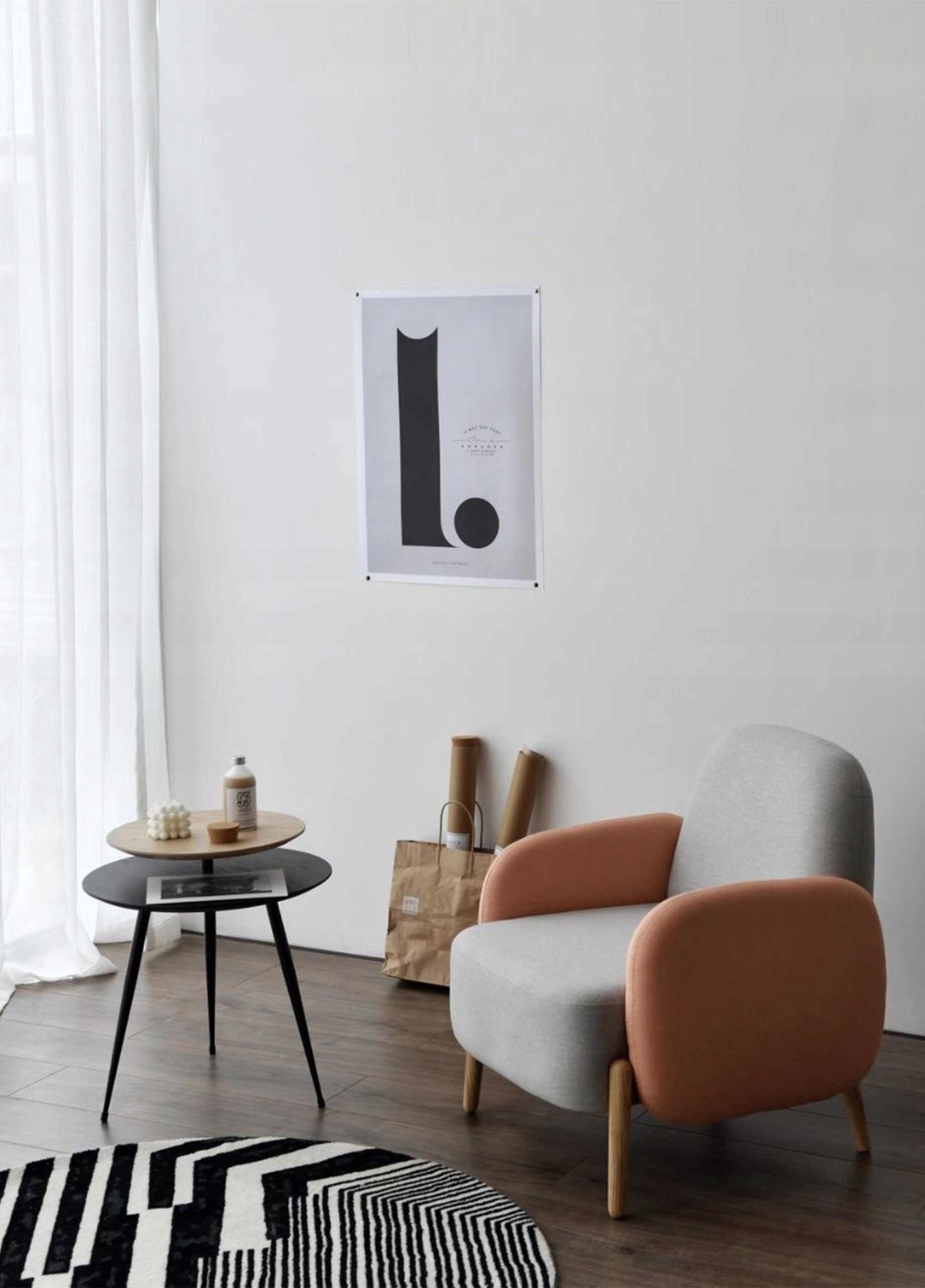 Wnętrze z jasnym fotelem o pomarańczowych podłokietnikach, obok którego stoi drewniany stolik kawowy z dwoma blatami. Na ścianie wisi plakat z dużą literą \'L\'.