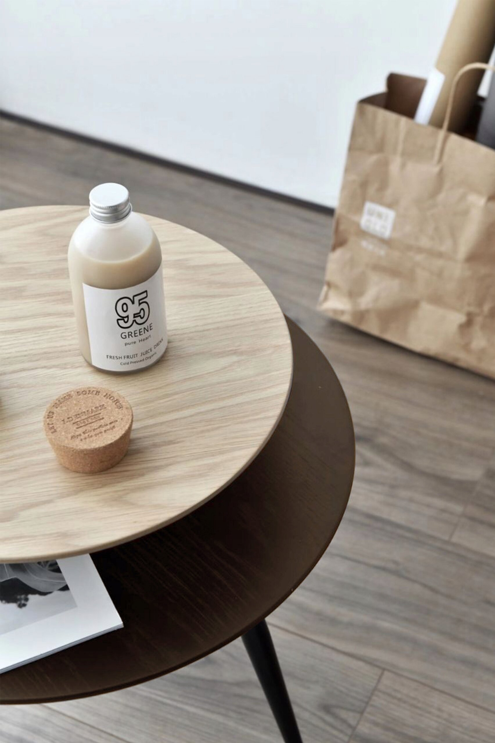 Na drewnianym stoliku kawowym z dwoma blatami znajduje się butelka z napisem \'95 GREENE\' oraz korek z tekstem. W tle widoczna jest papierowa torba stojąca na podłodze.