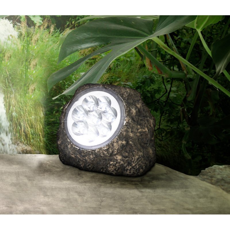 Lampa solarna w kształcie kamienia z widocznymi diodami LED na tle zielonej roślinności. Powierzchnia lampy imituje naturalny kamień.