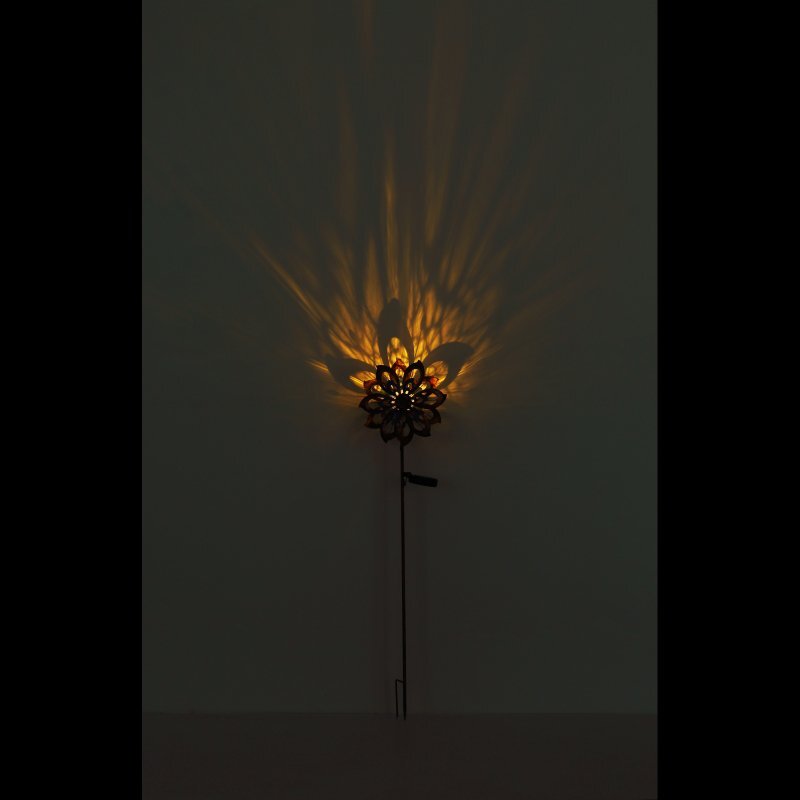 Lampa solarna LED w kształcie koła wiatrowego emituje ciepłe, żółte światło, tworząc wzór na ścianie. Lampa jest zamontowana na cienkim pręcie.