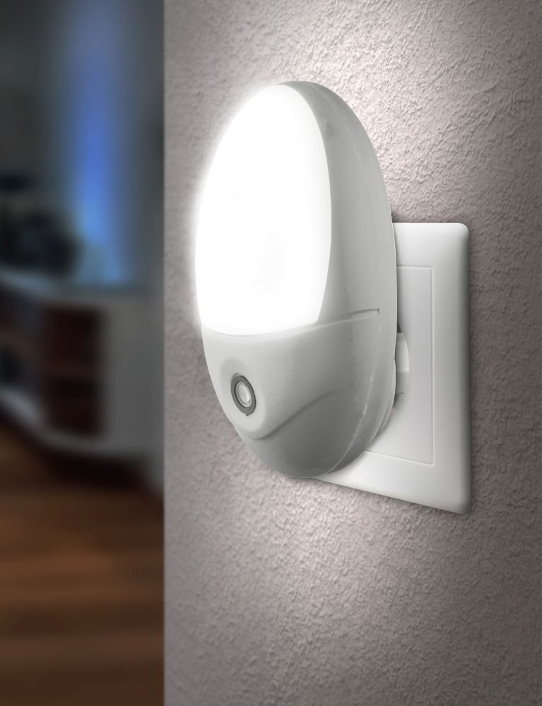 Biała lampka LED wtykowa z czujnikiem zmierzchu jest podłączona do gniazdka elektrycznego na ścianie. Emituje jasne światło, a na jej powierzchni widoczny jest okrągły czujnik.